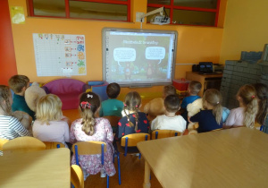 Grupa dzieci siedzi na krzesełkach przed tablicą interaktywną, oglądają bajkę o misiach.